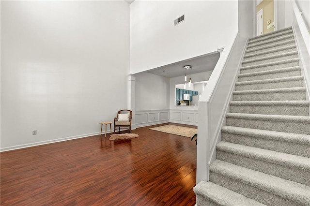 stairs with decorative columns, visible vents, a towering ceiling, wood finished floors, and a decorative wall