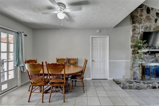 dining space with light tile patterned flooring, a stone fireplace, a textured ceiling, and ceiling fan