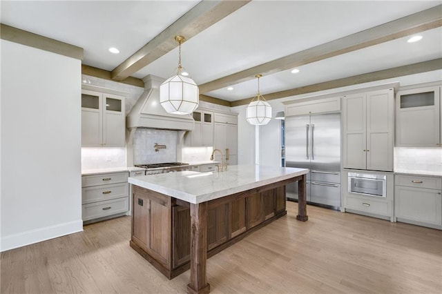 kitchen with stainless steel appliances, light hardwood / wood-style floors, custom range hood, an island with sink, and decorative light fixtures