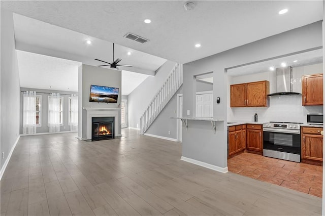 kitchen featuring wall chimney exhaust hood, appliances with stainless steel finishes, a peninsula, brown cabinetry, and open floor plan