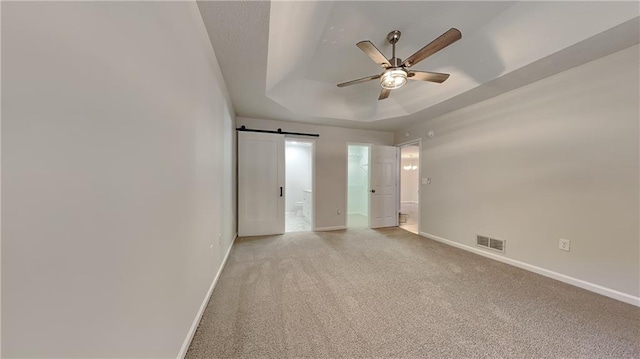 unfurnished bedroom with ensuite bathroom, light colored carpet, a raised ceiling, ceiling fan, and a barn door