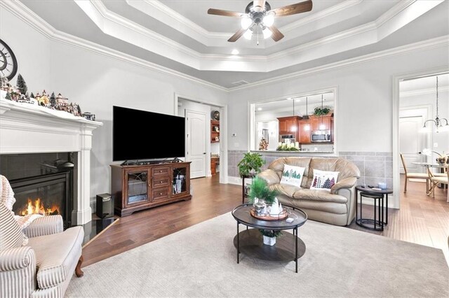 living room with a raised ceiling, wood-type flooring, a fireplace, ceiling fan with notable chandelier, and ornamental molding