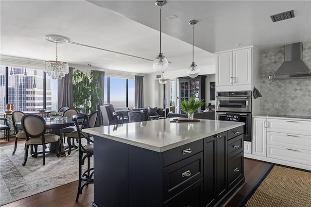 kitchen featuring white cabinetry, stainless steel double oven, wall chimney exhaust hood, and hanging light fixtures