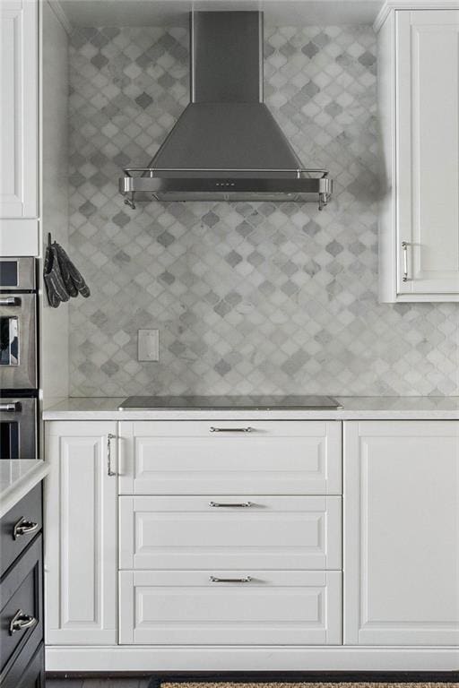 kitchen featuring double oven, black electric stovetop, white cabinets, decorative backsplash, and wall chimney exhaust hood