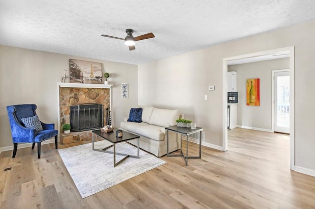 living room featuring ceiling fan, a stone fireplace, a textured ceiling, and light wood-type flooring