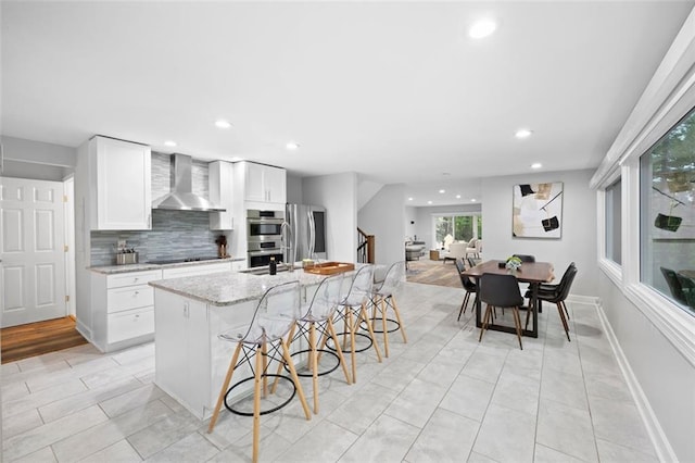 kitchen featuring wall chimney exhaust hood, white cabinetry, stainless steel appliances, and a center island with sink