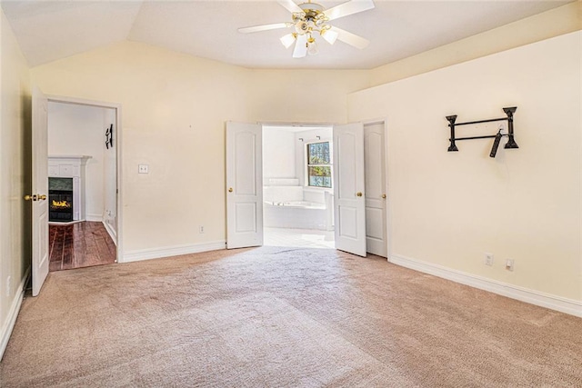 carpeted empty room featuring a warm lit fireplace, ceiling fan, lofted ceiling, and baseboards