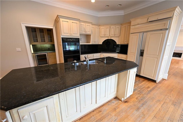 kitchen with sink, double oven, dark stone counters, light hardwood / wood-style floors, and a kitchen island with sink