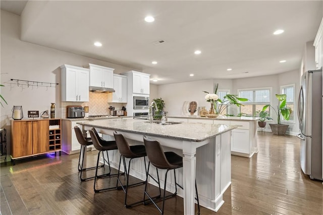 kitchen with white cabinets, dark wood-style floors, appliances with stainless steel finishes, light stone countertops, and a kitchen island with sink