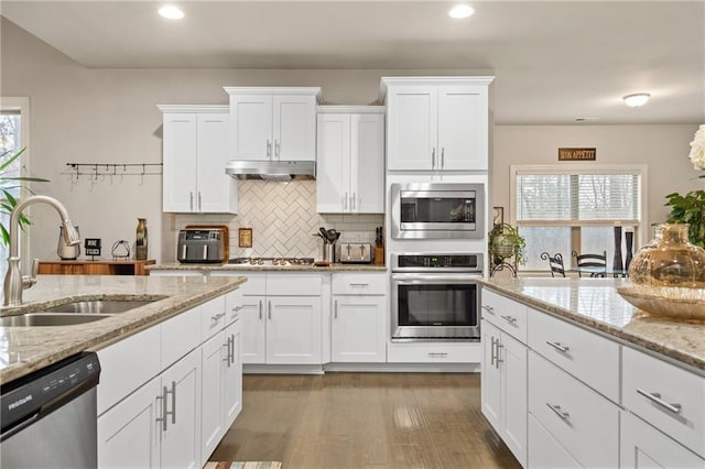 kitchen featuring under cabinet range hood, stainless steel appliances, a sink, white cabinetry, and light stone countertops