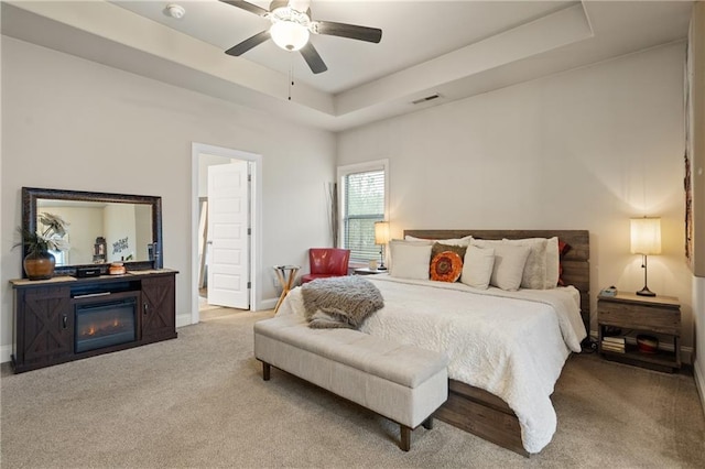 carpeted bedroom with a tray ceiling, a glass covered fireplace, a ceiling fan, and baseboards