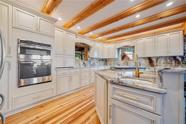 kitchen with light stone counters, beamed ceiling, tasteful backsplash, stainless steel double oven, and light wood-type flooring