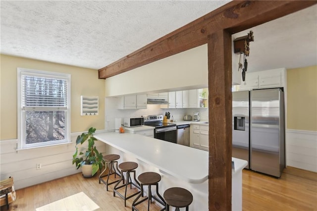 kitchen with under cabinet range hood, a textured ceiling, appliances with stainless steel finishes, a breakfast bar area, and wainscoting