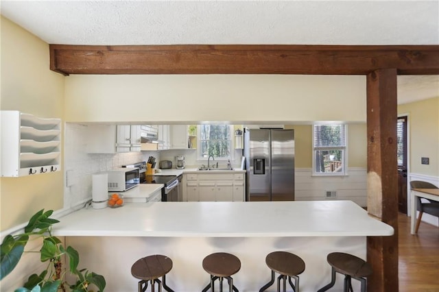 kitchen with a wainscoted wall, beamed ceiling, a sink, a peninsula, and appliances with stainless steel finishes