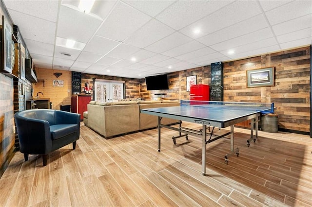 game room with wooden walls, light wood finished floors, and a drop ceiling