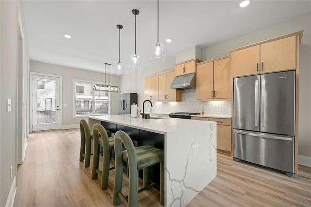 kitchen featuring stainless steel fridge, light brown cabinetry, an island with sink, and light stone countertops