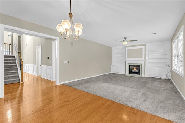 unfurnished living room with stairs, built in shelves, wood finished floors, and ceiling fan with notable chandelier