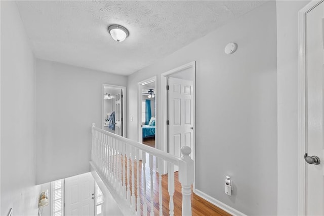 corridor featuring a textured ceiling, wood finished floors, an upstairs landing, and baseboards