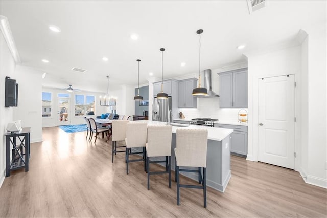 kitchen with gray cabinetry, decorative light fixtures, a center island with sink, appliances with stainless steel finishes, and wall chimney range hood