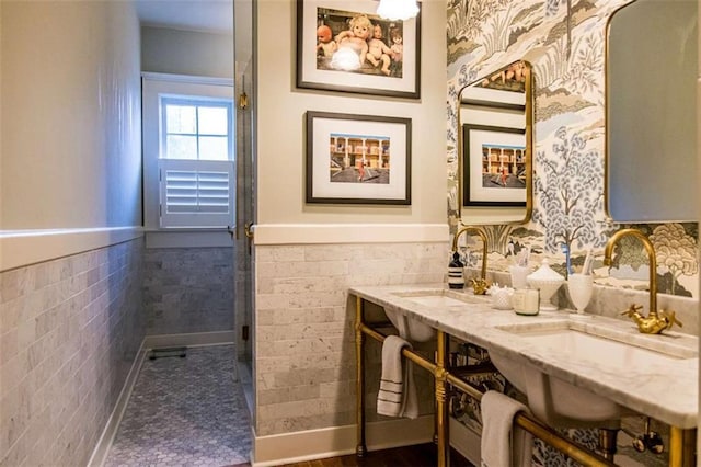 full bath featuring dual sinks, tile walls, wainscoting, and wallpapered walls