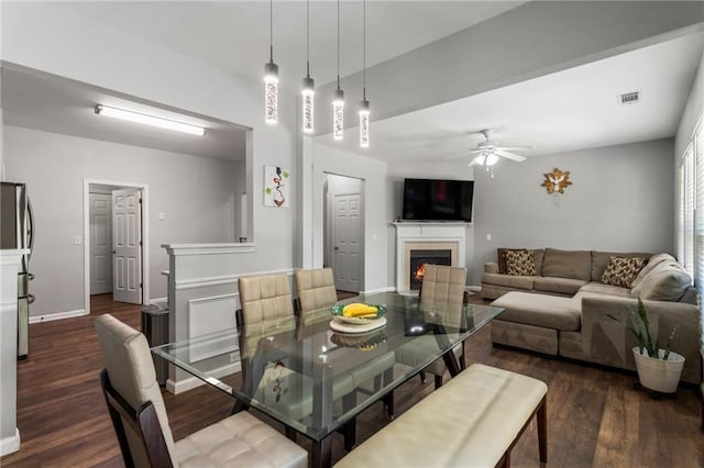dining room featuring baseboards, a ceiling fan, dark wood finished floors, and a warm lit fireplace