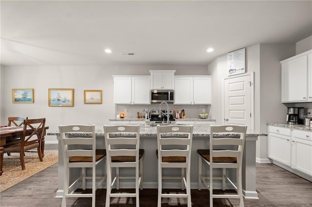 kitchen with white cabinets, an island with sink, dark wood-type flooring, stainless steel appliances, and light stone countertops