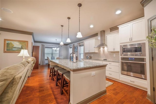 kitchen featuring decorative light fixtures, white cabinets, stainless steel appliances, a center island with sink, and wall chimney exhaust hood