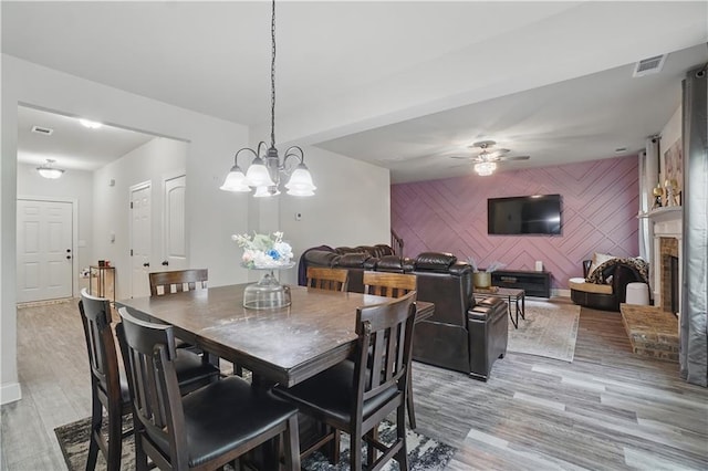 dining room featuring a ceiling fan, visible vents, a brick fireplace, an accent wall, and light wood-style flooring
