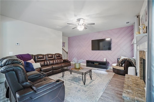 living area featuring visible vents, a glass covered fireplace, wood finished floors, an accent wall, and a ceiling fan