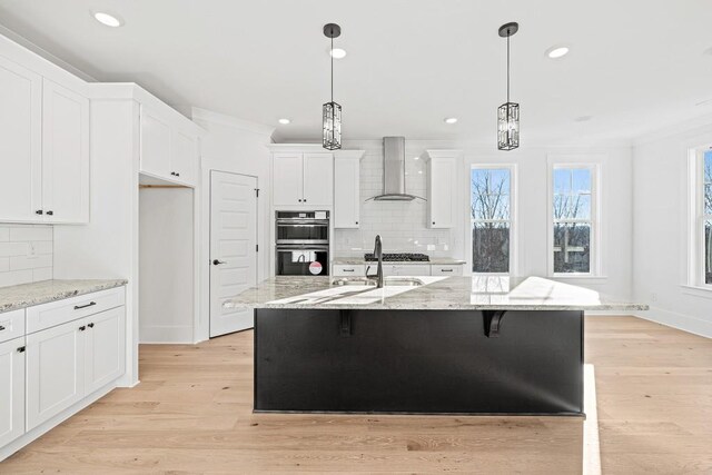 kitchen featuring double oven, pendant lighting, wall chimney exhaust hood, and a kitchen island with sink