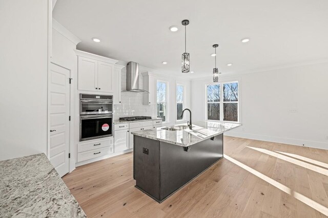 kitchen with stainless steel appliances, light stone counters, wall chimney range hood, sink, and white cabinetry