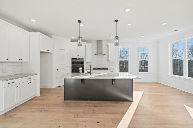 kitchen with a center island with sink, light stone counters, decorative light fixtures, wall chimney range hood, and white cabinets