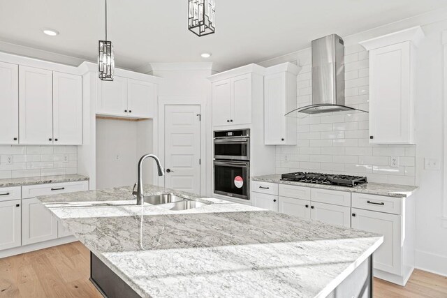 kitchen featuring a center island with sink, wall chimney exhaust hood, decorative light fixtures, black gas cooktop, and double oven