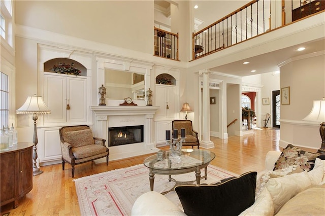 living area featuring crown molding, decorative columns, light wood finished floors, a towering ceiling, and a lit fireplace