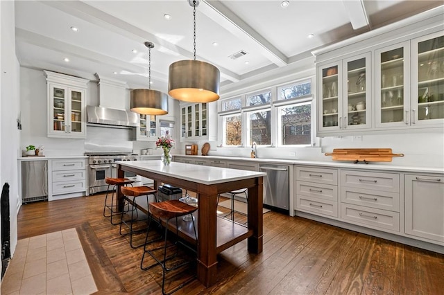 kitchen featuring beamed ceiling, stainless steel appliances, wall chimney exhaust hood, light countertops, and dark wood-style flooring