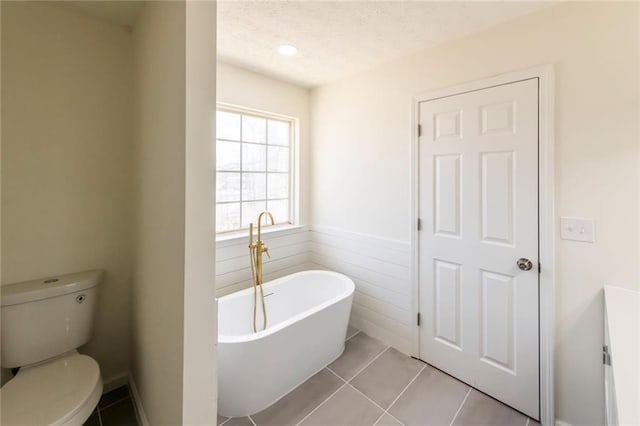 full bath with toilet, a wainscoted wall, tile patterned floors, a freestanding bath, and a textured ceiling