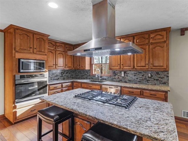 kitchen featuring brown cabinets, island range hood, a sink, light wood-style floors, and appliances with stainless steel finishes