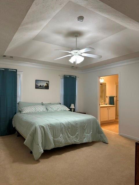 bedroom featuring connected bathroom, light carpet, ornamental molding, a raised ceiling, and ceiling fan