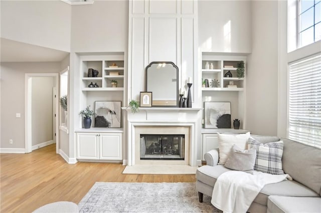 living room with built in shelves, baseboards, light wood-type flooring, a towering ceiling, and a glass covered fireplace