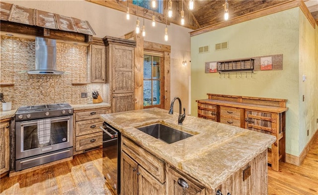 kitchen with appliances with stainless steel finishes, an island with sink, sink, wall chimney range hood, and light wood-type flooring