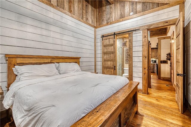 bedroom featuring a barn door, wooden walls, ensuite bath, and light wood-type flooring