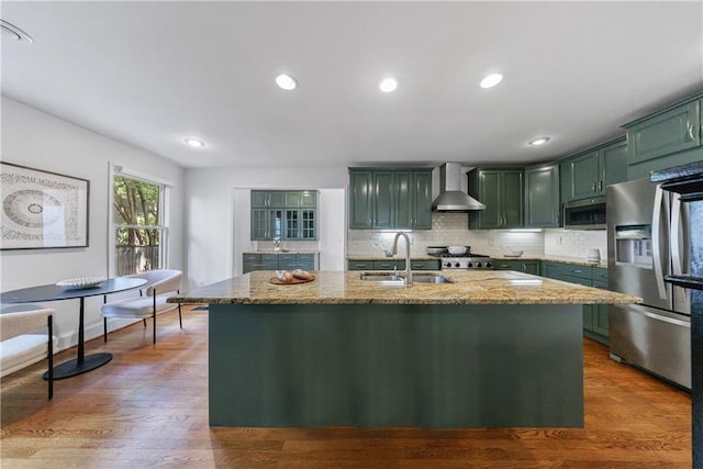 kitchen featuring light stone countertops, green cabinetry, stainless steel appliances, a sink, and wall chimney exhaust hood