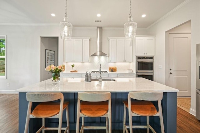 kitchen featuring wall chimney range hood, a kitchen island with sink, stainless steel double oven, a sink, and white cabinets