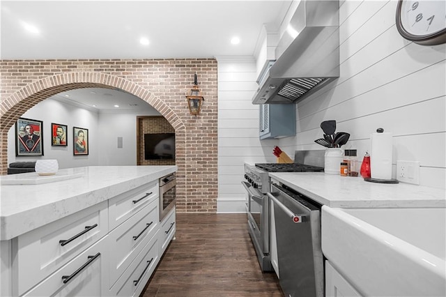 kitchen with brick wall, stainless steel appliances, white cabinets, wall chimney exhaust hood, and dark wood finished floors