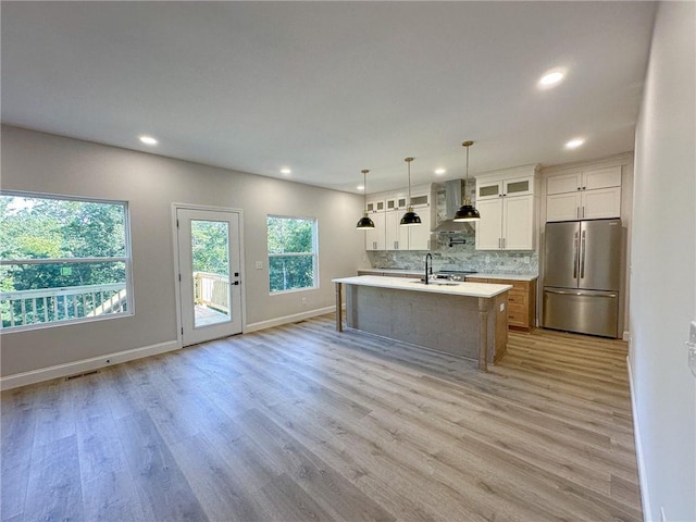 kitchen featuring light wood-type flooring, stainless steel appliances, wall chimney range hood, white cabinetry, and an island with sink