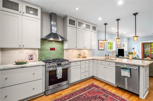 kitchen with a peninsula, wall chimney exhaust hood, a sink, light wood-style flooring, and appliances with stainless steel finishes