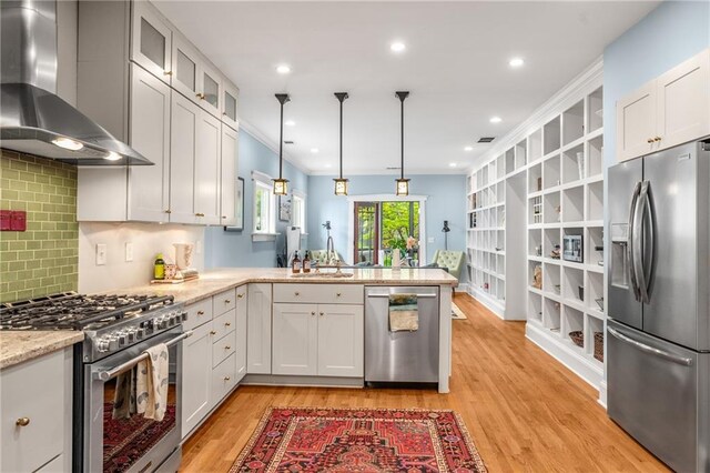 kitchen featuring wall chimney range hood, appliances with stainless steel finishes, a sink, light wood finished floors, and a peninsula