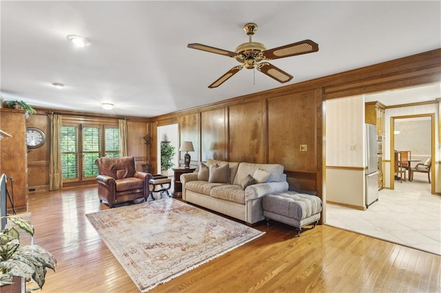living room featuring ceiling fan, light wood-type flooring, ornamental molding, wooden walls, and baseboards