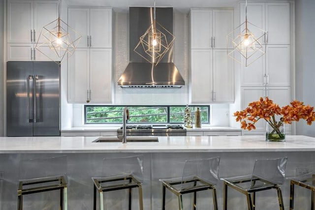 kitchen featuring high end refrigerator, wall chimney range hood, sink, hanging light fixtures, and light stone countertops