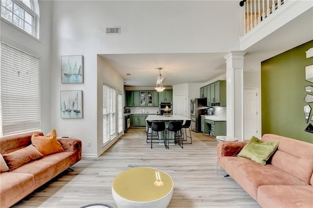 living area featuring baseboards, light wood finished floors, visible vents, ornate columns, and a high ceiling